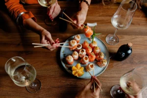 Varias personas compartiendo piezas de sushi y brindando con copas de vino de Jerez en una mesa de madera. Esta imagen se utiliza para ilustrar una entrada sobre platos internacionales que maridan a la perfección con los vinos de Jerez.